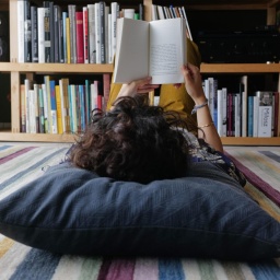 Eine junge Person liegt auf dem Boden und liest ein Buch. Der Kopf ist nur von hinten zu sehen und liegt auf einem großen grauen Kissen. 