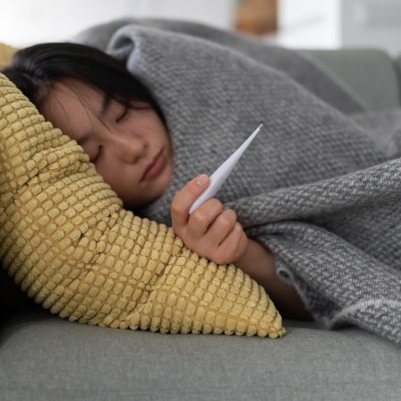 Eine Frau liegt eingemümmelt auf dem Sofa, sie hält ein Fieberthermometer in der Hand, sie ist krank