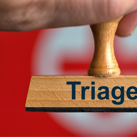 Ein symbolischer Holzstempel mit der Aufschrift "Triage", gehalten von einer Hand im Anschnitt vor einem unscharfen Hintergrund mit einem roten Kreuz. Foto mit Composing als Symbolbild.