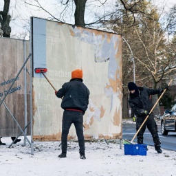 Zwei Männer mit bereiten eine leere Plakatwand auf einem verschneiten Straßenmittelstreifen vor für ein Wahlkampf-Plakat.