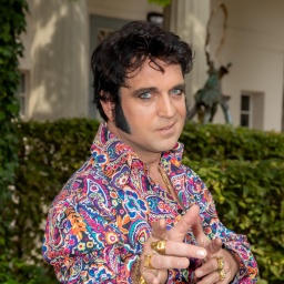 Nils Strassburg als Elvis in einem bunten Hemd und mit goldenen Ringen und schwarzen Koteletten. Er schaut direkt in die Kamera.