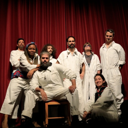 Die Band Wanderer Songs. Sechs Männer und zwei Frauen in weiß-brauen Overalls stehene vor einem roten Theatervorhang.