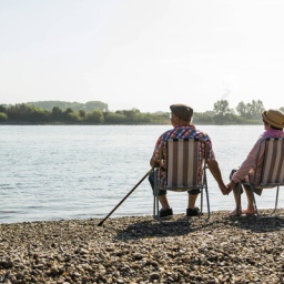 Ein älteres Paar sitzt Händchen haltend in Liegestühlen an einem Fluss.