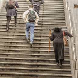 Eine ältere Dame mit Stock und ein junger Mann mit Kopfhörer gehen die Treppe einer U-Bahn herauf. Man sieht sie von hinten.