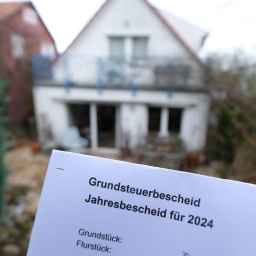 Ein Grundsteuerbescheid für 2024 wird vor einem Privatgrundstück hochgehalten