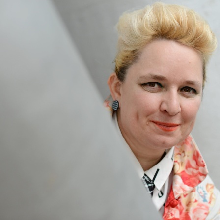 Eine Frau mit blondierten Haaren schaut um einen Vorsprung und lächelt in die Kamera. Es ist die Künstlerin Mariola Brillowska.
