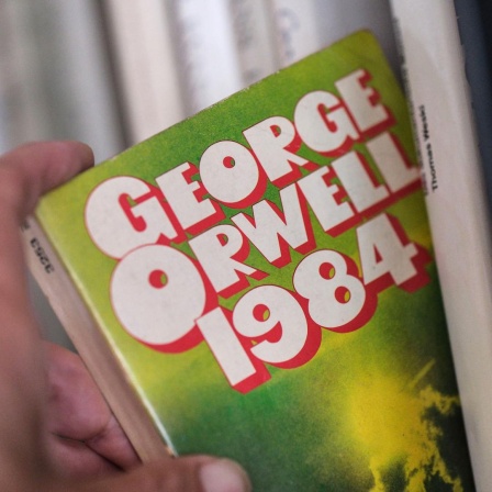 Ein ältere Ausgabe des Romans "1984" von George Orwell wird von einer Hand aus einem Bücherregal genommen. 
