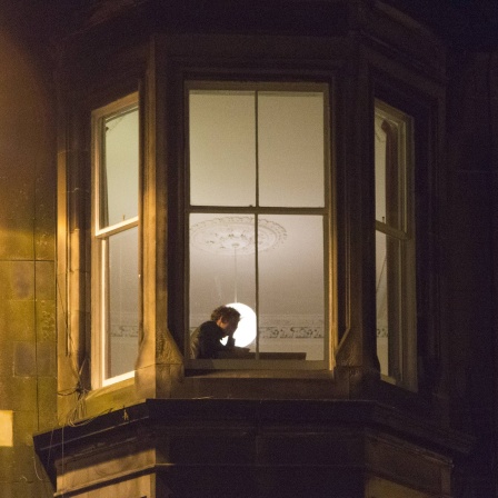 einsamer junger Mensch am Fenster, sitzt am Schreibtisch, stützt den Kopf in der Hand, von der Straße aus gesehen