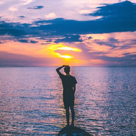 Ein Mann steht auf einem kleinen Felsen im Meer und schaut in die Abendsonne.