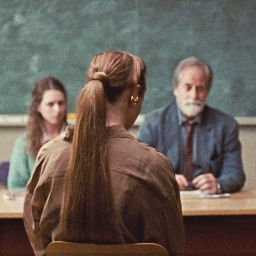 Eine Frau langem Pferdeschwanz sitzt in einem Klassenzimmer vor einem Pult, dahinter drei Personen - Szene aus "Armand"