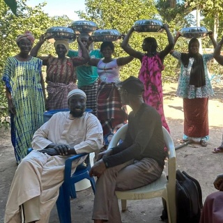 Männer sitzen auf Plastikstühlen, dahinter stehen Frauen mit Kopftüchern (Foto aus dem Senegal)