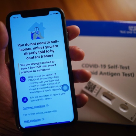 Auf einem Handydisplay ist die britische Corona-Warnapp zu sehen, im Hintergrund liegt ein Corona-Selbsttest.