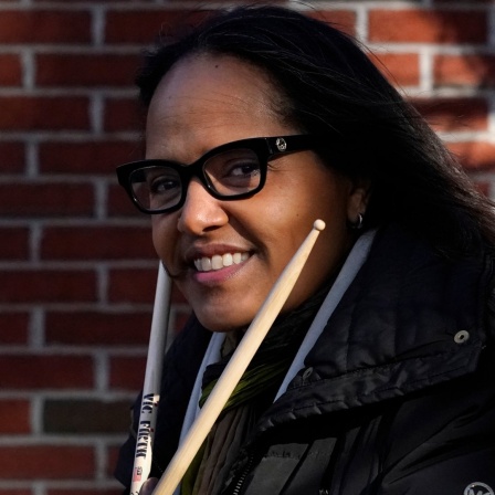 Terri Lyne Carrington lächelt in die Kamera und hält zwei Schlagzeugsticks in ihren Händen.