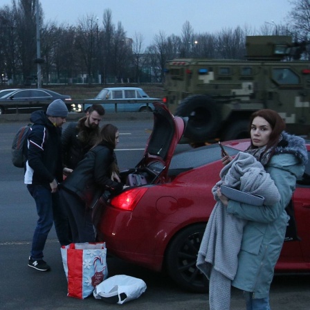 Menschen laden ihre Sachen in ein Auto, während ein Militärfahrzeug auf einer Straße in Kiew, Ukraine, vorbeifährt.