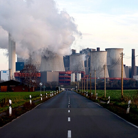 Blick auf eine leere Straße, dahinter das Kohlekraftwerk Niederaußem. Aus mehreren Schornsteinen steigt Rauch auf.