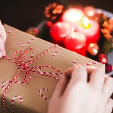 Eine Person verpackt ein Weihnachtsgeschenk an einem Tisch. Davor ist ein Adventskranz.