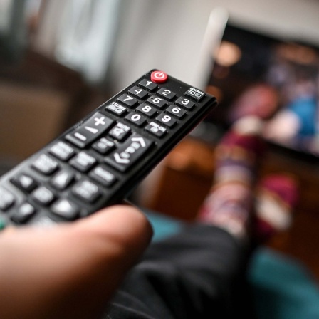 Ein Mensch sitzt mit einer Fernbedienung in der Hand vor einem Fernseher.