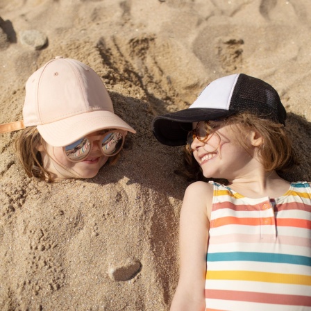 Zwei Kinder liegen nebeneinander am Strand und lachen sich an. Das linke Kind ist im Sand eingegraben. Nur der Kopf ist zu sehen.
