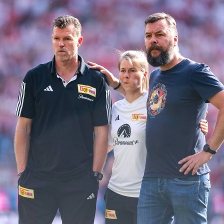 Union Berlins Trainer Marco Grote, Co-Trainerin Marie-Louise Eta und Geschäftsführer Kommunikation Christian Arbeit stehen Arm in Arm am Spielfeldrand bei der Partie in Köln. (Archivbild)