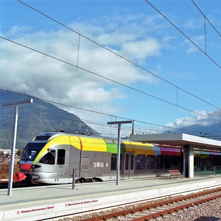 Ein Bahnhof in dem ein grauer Zug steht. Im Hintergrund ein Bergpanorama bei Sonnenschein.