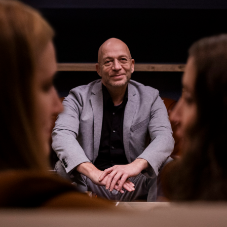 Blick auf Eric Hegmann, der auf einer Couch sitzt, im Vordergrund zwei Frauen, die sich angucken