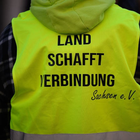 Ein Teilnehmer einer angemeldeten Demonstration steht vor dem Sächsischen Staatsministerium für Energie, Klimaschutz, Umwelt und Landwirtschaft mit einer Schutzweste mit dem Schriftzug „Land schafft Verbindung Sachsen e.V.&quot;.