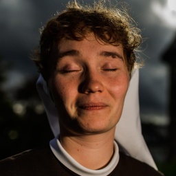 Eine junge Frau mit Nonnenschleier sitzt lächelnd und mit geschlossenen Augen in einem Garten. Hinter ihr die Umrisse eines Klostergebäudes.