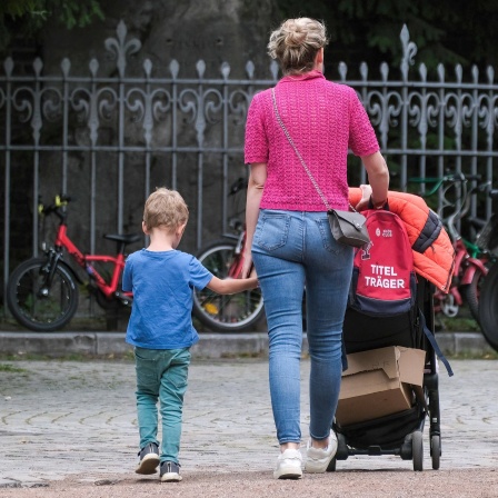 Eine Mutter hält ein Kind an der Hand und schiebt einen Kinderwagen.