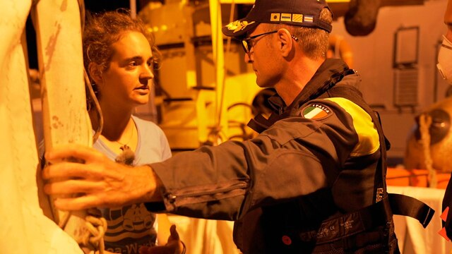 Carola Rackete spricht mit einem Mitarbeiter der "Guardia de Finanzia". Es ist Nacht und sie werden von gelbem Licht beleuchtet.
