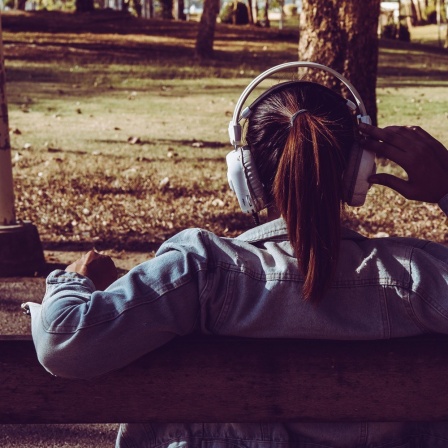 Die Parkbank ist eine Station der akustischen Reise. Eine junge Frau sitzt mit Kopfhörern auf einer Parkbank.