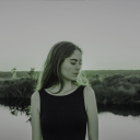 Eine junge Frau, mit dem Kopf zur Seite gesenkt, steht vor einem See. Im Hintergrund ist eine Wiesenlandschaft.