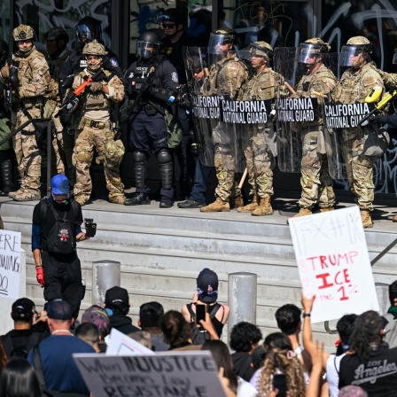 Menschen protestieren in Los Angeles, während Einsatzkräfte der Nationalgarde vor einem Regierungsgebäude stehen. 