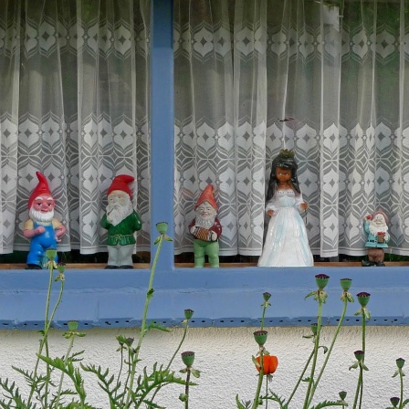 Schneewittchenfigur und sieben Gartenzwerge aufgereiht im Fenster eines Gartenhauses