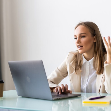 Eine junge Geschäftsfrau mit Blazer und weißer Bluse sitzt vor einem Laptop am Schreibtisch und scheint mit einer anderen Person auf dem Bildschirm zu sprechen.