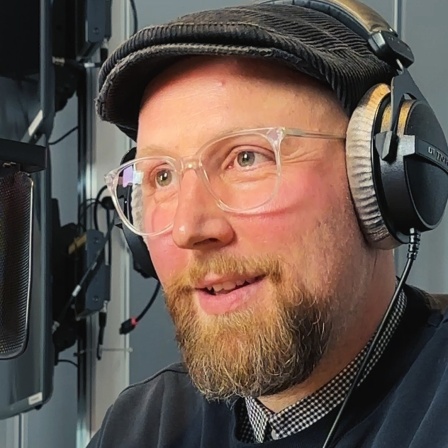 Ein Mann mit Bart, Brille und Schirmmütze hat Kopfhörer auf und spricht in ein Mikrophon. 