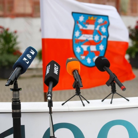 Die Mikrofone verschiedener Medienanstalten stehen auf einem Podest vor der thüringischen Landesflagge.