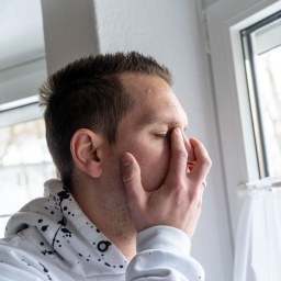 Ein junger Mann steht traurig und verzweifelt an einem Fenster, er hält sich das Gesicht vor Schmerz.