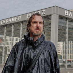 Ein Mann in grün gekleidet mit nassen Haaren steht im Regen an einer Berliner Kreuzung - Szene aus dme Film "Das Licht"