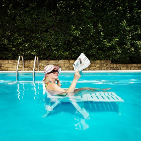 Eine Frau mit einem Sonnenhut liegt auf einer Sonneliege in einem gefüllten Pool und liest Zeitung.