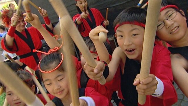 Eine Gruppe japanischer Kinder mit roten Stirnbändern und rot/schwarzen Karateanzügen mit langen Stöcken in den Händen.