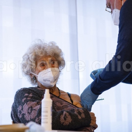 Eine ältere Dame mit grauen Locken und weißem Mundschutz hat den Ausschnitt ihres Pullovers bis über den linken Oberarm gezogen, so dass ein Arzt sie dort impfen kann. 