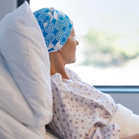 Eine junge Frau mit blauer Kopfbedeckung liegt nachdenklich im Krankenhausbett, hat die Hände ineinander gefaltet und schaut aus dem Fenster raus.