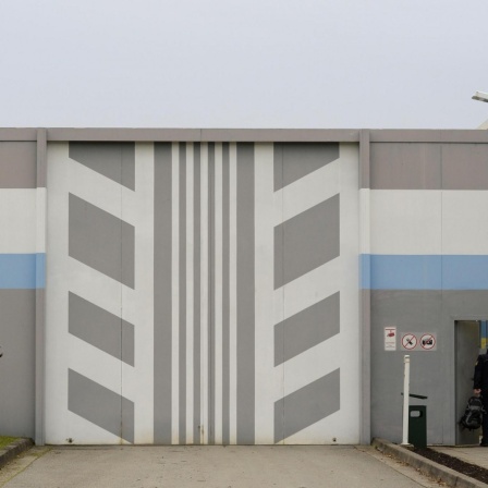 Ein Mann betritt die Jugendvollzugsanstalt Adelsheim in Baden-Württemberg. Das Rolltor hat ein grau-weißes Muster, die Mauer ist grau-weiß-blau gestrichen.