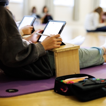 Eine Schülerin einer sechsten Klasse sitzt mit anderen Schülerinnen in einem Schulraum  auf dem Boden und arbeitet an ihrem Tablet.