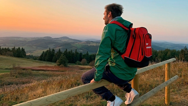 Daniel Aßmann sitzt auf einem Holzzaun und schaut auf den Sonnenuntergang über einer bewaldeten Hügellandschaft der Rhön