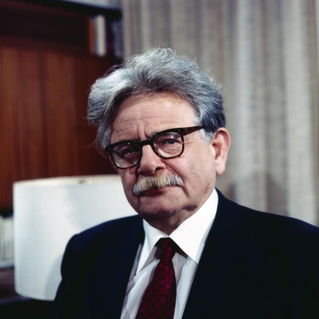 Porträt von Elias Canetti, der eine Brille mit dickem schwarzem Rand und einen schwarzen Anzug mit dunkelroter Krawatte trägt.