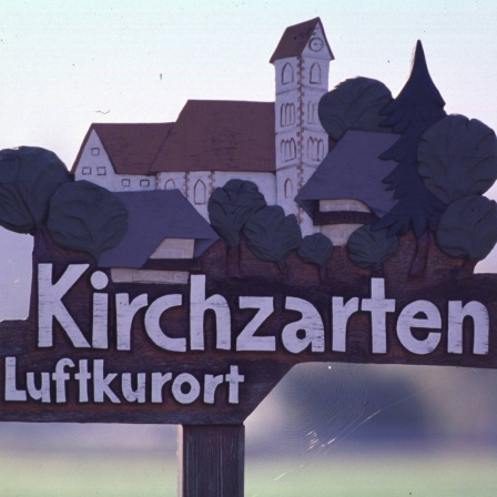 Auf dem Wegweiser zum Luftkurort Kirchzarten sind barocke Gebäude und grüne Bäume zu sehen.