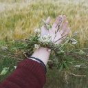 Ein ausgestreckter Arm mit einem Kranz aus wildblühenden Pflanzen um das Handgelenk vor einer Sommerwiese.