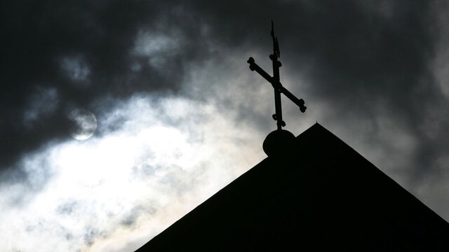 Im Gegenlicht und vor einem wolkigen Himmel ein Kreuz auf einer Kirche