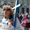 Eine Frau mit Sonnenbrille, mit halb offenem Mund und einem großen, blauen Kreuz in Ihrem Arm nimmt an einer Demonstration in Verona Teil.
